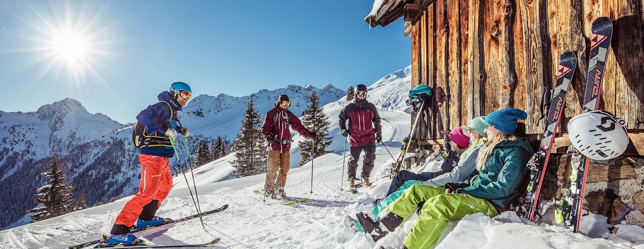 Skigruppe entspannt in der Sonne vor einer Almhütte in verschneiter Berglandschaft